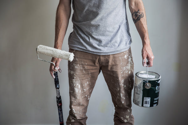 A mam holding a paint roller and paint can, standing in front of a white wall. 